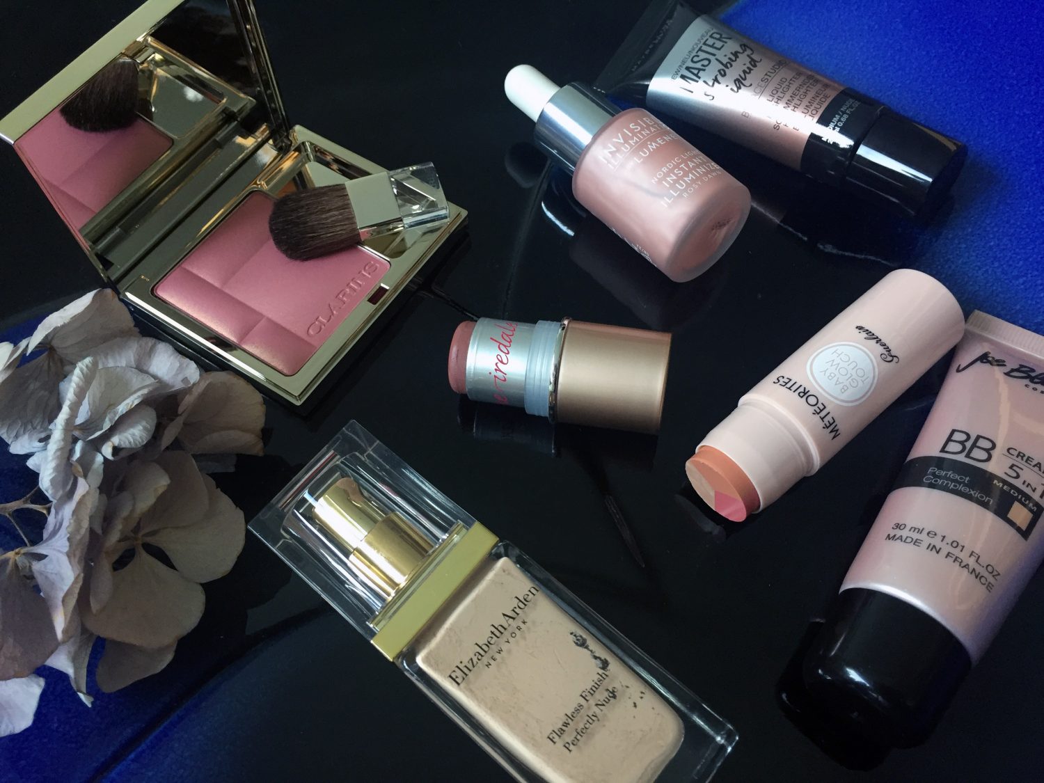 Vasemmalla Clarins Blush Prodige -poskipuna sävy 09, Elizabeth Arden Flawless Finish Perfeetly Nude Makeup spf 15 -meikkivoide, Jane Iredale In Touch Cream Blush -voideposkipuna sävy Candid, Lumene Invisible Illumination Rosy Dawn -valopisarat, Maybelline Master Strobing Liquid -korostusväri sävy medium/nude, Guerlain Météorites CC Luminizer Easy Strobing -poskipuna-korostus-varjostuspuikko ja Joe Blasco BB Cream 5 in 1 -voide väsy medium.